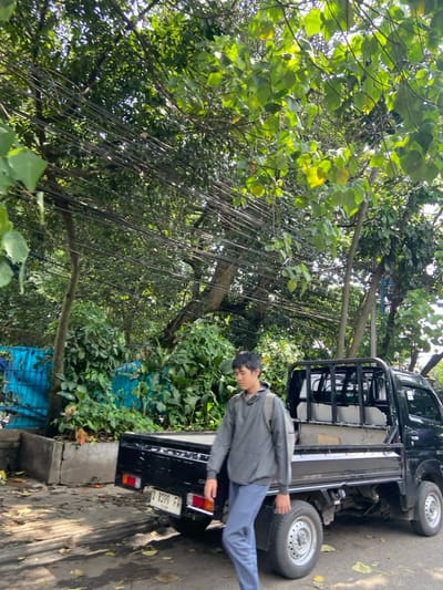 A young man is standing next to a black pickup truck parked on a paved road in a dense, overgrown area. The man is wearing a grey hoodie and blue jeans and has short, dark hair. The truck is parked with the bed facing the road, and the cab is facing the trees. The man is looking down at the ground, and his right hand is resting on the side of the truck. The truck has a license plate that reads "D 8399 FV." There are many trees and shrubs surrounding the road and truck, and the ground is covered with dry leaves and dirt. The location is in Kecamatan Cimahi Selatan, Indonesia, at latitude -6.904 and longitude 107.565. There are many power lines running through the trees and shrubs, but no other people are visible.