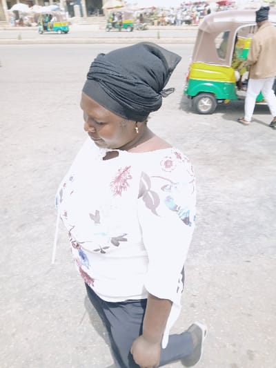 The scene is set in Masvingo, Zimbabwe, depicting an outdoor urban environment under bright daylight. In the foreground, a woman, appearing middle-aged, is captured from the chest down to the upper thigh, looking slightly downward and to her left. She wears a black headwrap, a white long-sleeved blouse adorned with purple, blue, green, and brown floral patterns, dark-colored trousers, and dark sneakers with white soles. A single gold-colored hoop earring is visible in her right ear. Her right arm is bent at the elbow, with her hand resting on her upper right thigh.

The background features a paved street or open area. To the right, a brown, green, and yellow three-wheeled auto-rickshaw type vehicle is visible. A person, clad in a light brown jacket, white trousers, and a dark head covering, walks near this vehicle. Across the street, in the mid-ground, at least two other three-wheeled vehicles are visible, one green and yellow, another light blue and yellow. Beyond the street, indistinct light-colored buildings and multiple pedestrians populate the far background, indicating a busy commercial or public area.