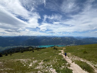 A man walks along a gravel path on a grassy mountainside with a wide view of mountains and a turquoise lake in the distance. The man, wearing a blue shirt and black shorts, is walking away from the camera. The path curves to the right and down into the valley. The mountains in the background are green and layered, with a bright turquoise lake in the middle ground. The sky is partly cloudy with dramatic white clouds against a blue sky. In the foreground, the grass is green with scattered rocks. The scene appears to be in the area around Sankt Gilgen, Austria, known for its alpine scenery and lakes.