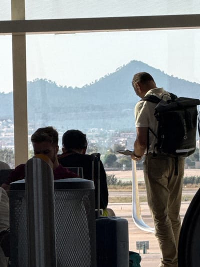 The image depicts a group of people in what appears to be an airport or train station waiting area, likely in el Prat de Llobregat, Spain, given the context. The perspective is unusual, as the scene is captured sideways, suggesting the photo was taken with the device oriented vertically and then rotated.
In the foreground, a man wearing a light-colored t-shirt and khaki pants is visible. He has a dark backpack on his shoulders, with "Earth-Pak" printed on it. He is holding a white object, possibly a boarding pass or ticket, in his hand. His head is angled downwards, and his expression is not clearly discernible.
Behind him, other individuals are seated, though their features are less clear due to the angle and lighting. The scene is characterized by a large window offering a view of a hazy landscape with hills and some buildings. The natural light suggests it is daytime, possibly late afternoon given the warm tones.
The waiting area itself has metallic seating and railings, indicative of a public transportation hub. The overall atmosphere seems calm, with people waiting for their departure. The orientation of the photo and the slightly blurred background create a sense of movement or perhaps a candid snapshot. No specific weather conditions are evident, other than the hazy appearance of the distant landscape.