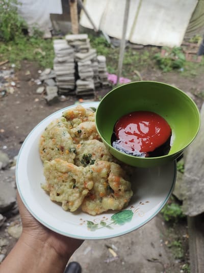 A hand holds a white plate with a light blue rim, on which rests a serving of fritters. The fritters appear to be made with finely chopped vegetables, possibly carrots and greens, mixed into a batter and fried. They are golden brown and irregular in shape. To the right of the fritters, a green bowl contains a dollop of red sauce, likely chili sauce or ketchup, with a darker sauce, possibly soy sauce, pooled beneath it. The plate and bowl are positioned in the foreground, with a slightly blurred background of an outdoor environment. This environment includes patchy green vegetation, piles of what appear to be concrete or brick rubble, and a weathered white tarp or sheeting. The overall impression is of a casual, possibly rustic, outdoor setting.
