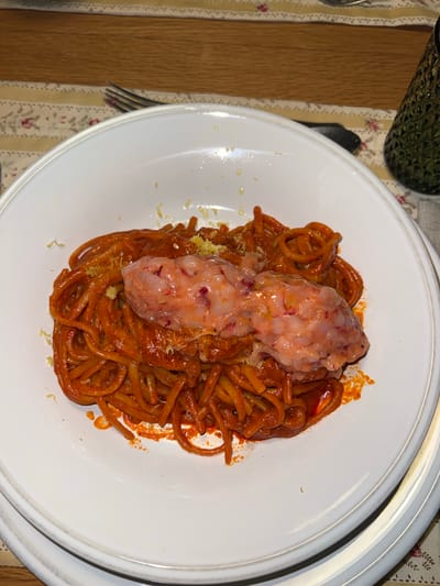 A top-down view depicts a white plate containing spaghetti with a red sauce, topped with a pale, irregularly-shaped mass. The plate is positioned on a tablecloth with a floral pattern, adjacent to a wooden table surface and a dark green glass object. A fork lies to the left of the plate. The scene is located in Gondarém, Portugal.