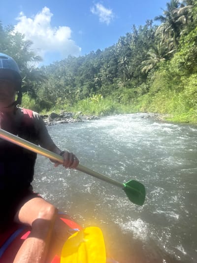This is a first-person perspective image taken from a raft on a river in Sidemen, Indonesia. The person in the raft is wearing a blue helmet and a dark shirt with a red and gray vest. Their hand is holding a paddle with a metallic shaft and a green paddle blade, which is extended into the turbulent, whitewater river. The raft itself is partially visible at the bottom of the frame, appearing to be red and blue with a yellow object on the right. The riverbanks are densely covered with lush green vegetation, including tall grasses and palm trees, indicative of a tropical environment. The sky above is blue with scattered white clouds. The overall scene depicts an outdoor adventure activity, likely white-water rafting, in a scenic, natural setting.