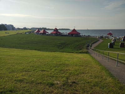 The image depicts the Cuxhaven Wadden Sea National Park, a coastal area in Germany. In the foreground, a lush green lawn slopes gently towards a paved walkway bordered by a metal railing. Several people are walking along the path. In the midground, a row of red-roofed buildings with conical tops lines the waterfront. Beyond these buildings, the blue sea stretches to the horizon, with a distant lighthouse visible on the left. The sky is overcast with wispy clouds. The scene suggests a recreational area with amenities for visitors enjoying the coastal environment.