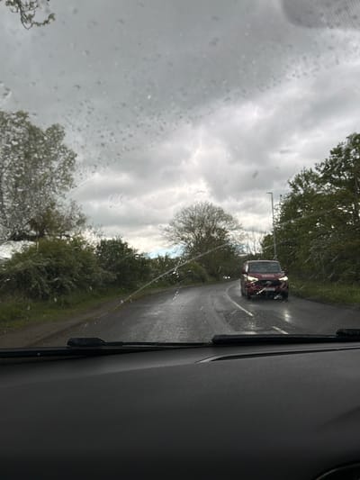 The image depicts a view from inside a vehicle, likely a car, on a rainy day in what appears to be a rural area. Through the rain-streaked windshield, a road is visible, with a red car approaching in the distance. The road is flanked by trees and greenery. The sky is overcast with heavy clouds. The interior of the vehicle is visible on the right side of the frame, showing part of the dashboard and door. There are no people visible in the scene.
Specific to the request, the location in the city of Durham, United Kingdom, cannot be specifically confirmed from the image alone; however, the rural road and vegetation are reminiscent of areas outside the city center. It would require additional information or landmarks to definitively pinpoint the location.