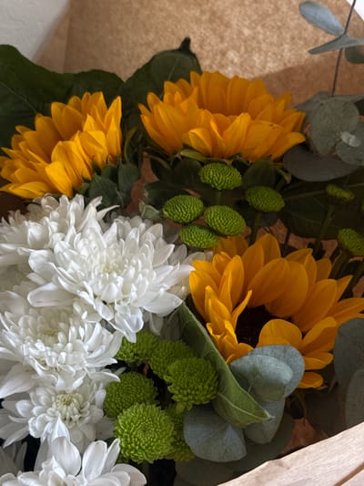This is a close-up, eye-level, medium shot of a bouquet of flowers. The bouquet features several bright yellow sunflowers with dark centers, mixed with a cluster of fluffy white chrysanthemums. Small, bright green button-like flowers are interspersed throughout the arrangement, as are several sprigs of dusty green eucalyptus leaves. The flowers are presented in a light brown paper wrapping. The background is softly blurred and appears to be a textured brown surface, possibly cardboard. The lighting is bright and natural, highlighting the vibrant colors of the flowers. There are no people or specific locations visible in the image; it is a still life of a floral arrangement.