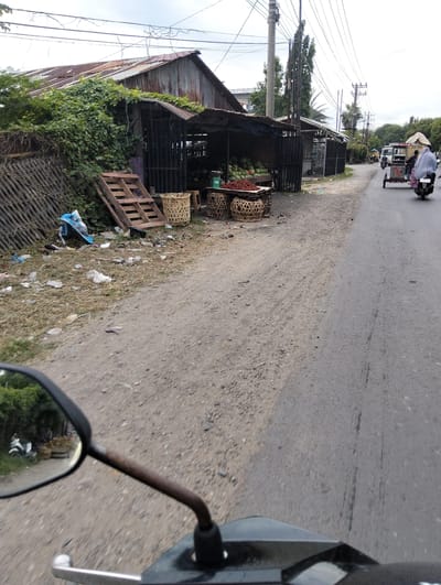 The image depicts a roadside scene in Tambonbaroh, Indonesia, featuring a fruit stand with a selection of watermelons and rambutan. A rustic wooden stall with a corrugated metal roof displays the produce. Baskets of fruit are prominent, with a scale visible to the right. The surrounding area is unpaved, with debris and vegetation present. In the background, a road runs parallel to the stall, with a motorbike carrying passengers in transit. Power lines crisscross the sky, and several trees are visible. The overall impression is one of a typical street-side market in a developing urban or semi-urban environment.
