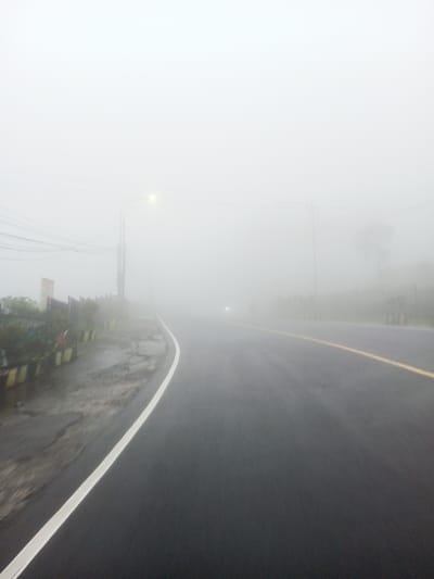 This image depicts a section of a two-lane asphalt road in Dawathang_Dorjibi_ Kashingtsawa, Bhutan, under conditions of dense fog. The road surface appears wet, suggesting recent precipitation or high humidity. A solid white line delineates the left edge of the road, while a solid yellow line marks the approximate center or right edge of the visible lane, both receding into the extreme low visibility caused by the fog. On the left side of the road, a utility pole with an illuminated street light emits a faint yellow glow, partially piercing the fog. Multiple overhead utility wires extend horizontally and diagonally from the pole. Below the wires, a red and white sign is partially visible amidst vegetation and what appears to be a fence or barrier. Further along the right side of the road, several roadside markers or small barriers with yellow tops are discernible, also fading into the fog. In the far distance, approximately centered on the road, a faint light source, consistent with a vehicle headlight, is visible through the thick fog. No people or vehicles are clearly visible aside from the distant light source. The environment is characterized by heavy atmospheric obscuration, rendering distant details indistinct.