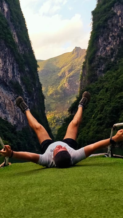 A man lies supine on a green surface, legs raised, arms extended. The scene is framed by steep, tree-covered cliffs typical of the Đồng Văn Commune in Vietnam. A landscape with verdant hills and a small settlement is visible in the distance between the cliffs, beneath a partly cloudy sky. The man wears dark shorts, a white t-shirt, shoes with dark socks, and a black wristband. He appears relaxed, with his arms outstretched. A metallic bar and rope are near his hands.