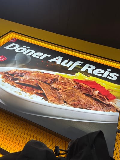 An illuminated, rectangular sign displaying a food advertisement is the central subject of this image, captured at a diagonal angle in what appears to be an indoor setting in Kaiserslautern, Germany.
The sign prominently features the German text "Döner Auf Reis" in large white letters on a dark background. Below the text, a vibrant image of the dish "Döner on Rice" is shown in a white bowl. The dish consists of generous servings of thinly sliced, seasoned meat (Döner), white rice, and bright red elements (likely chopped tomatoes or peppers), along with some yellow components, which appear to be french fries, arranged on top. Small, reddish star-shaped graphic elements are visible around the text, and small red icons resembling a "thumbs up" or "like" button are also present at the bottom left of the sign.
The sign is bordered by a distinctive yellow frame with a reflective, fine diamond or mesh pattern. Beyond this frame, the immediate background is a dark, solid surface, while on the far right, a yellow wall with the same diamond pattern is visible. Part of a person, identifiable by a dark sleeve or hoodie, is partially obscuring the right edge of the sign and the adjacent wall. The strong illumination from the sign itself contrasts with the darker surrounding environment, suggesting it might be evening or an interior with focused lighting.
