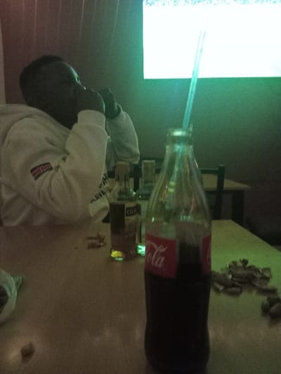 A person in a white sweater with a Kenyan flag patch drinks a Coca-Cola bottle using a straw. The bottle is tilted on a table, and several small objects are scattered nearby. A bright screen glows in the background.