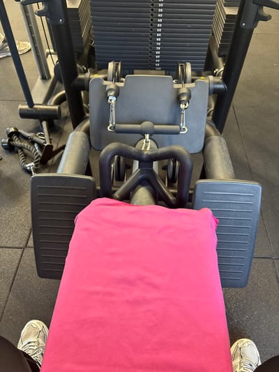 This image, captured from a top-down perspective, shows a weight training machine in an indoor gym setting in Córdoba, Spain. The primary subjects are the gym equipment and the feet of an individual wearing sneakers.

The machine features a substantial weight stack on the left side, with visible resistance levels numerically marked from 30 to 100 (specifically: 30, 35, 40, 45, 50, 55, 60, 65, 70, 75, 80, 85, 90, 95, 100). The machine itself is robust, constructed from dark grey and black metal, with a cable and pulley system evident. Two textured footrests are visible, suggesting the machine might be a seated row or similar lower body/back exercise equipment.

A bright pink towel is neatly draped over the padded seat or support area of the machine, indicating that the equipment is either in use, has just been used, or is prepared for use. Two feet, clad in white and grey athletic sneakers, are visible on the black rubber mat flooring – one on the bottom right and another partially seen on the bottom left, positioned near the machine.

The scene suggests an ongoing or recently concluded workout session. The gym floor is covered with standard black interlocking rubber mats. No specific time of day, weather, or emotions are discernible, but the functional setup points to a typical fitness environment.