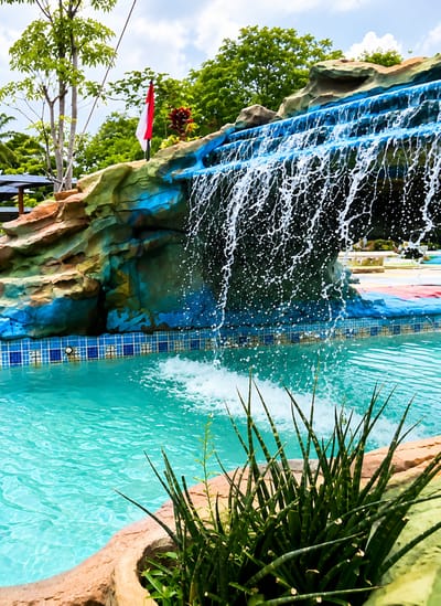 An outdoor artificial waterfall cascades into a swimming pool, with lush green trees and a cloudy blue sky visible in the background. The waterfall is designed to resemble a rock formation, painted in shades of blue and brown. A red and white Indonesian flag is planted on top of the rock formation. In the foreground, a cluster of spiky green plants grows out of a planter at the edge of the pool. The swimming pool itself is a bright turquoise color, with a tiled border featuring a blue and white checkered pattern. The scene appears to be set in a resort or water park. The exact location is identified as Eampat Kwalamencirim, Indonesia.