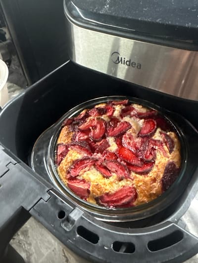 A strawberry cake is baked inside a glass dish, which is placed in the basket of a Midea air fryer. The cake is golden brown with visible slices of strawberries on top. The air fryer is black with a silver metallic band featuring the "Midea" logo. The scene is indoors and appears to be a kitchen. The specific location in Chakvi, Georgia, is not determinable from the image.