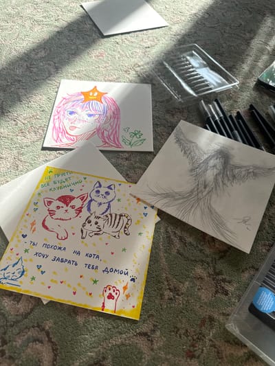 The image depicts a collection of hand-drawn illustrations and art supplies scattered on what appears to be a carpeted surface. There is a drawing of a pair of wings done in pencil, a colorful drawing of a person with pink hair and an orange star, and a drawing of three cats with text in Russian. The text in Russian says "Ты похожа на кота, хочу забрать тебя домой", which translates to "You look like a cat, I want to take you home". There is also a plastic container and a set of drawing pens nearby. There is no mention or indication that this image depicts Uryupinsk, Russia.
