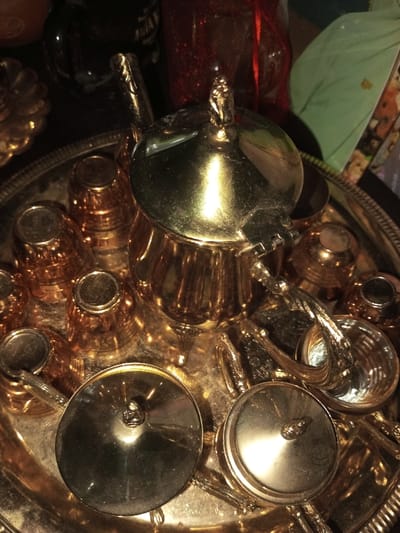 A shiny, gold-colored tea set sits on a silver tray. The set includes a teapot with a hinged lid and ornate handle, several small cups with vertical ribbing, and lidded sugar bowls. The reflective surfaces create bright highlights. The tea set is surrounded by various objects, including a dark bottle and a decorative red container. A green object is visible in the background.