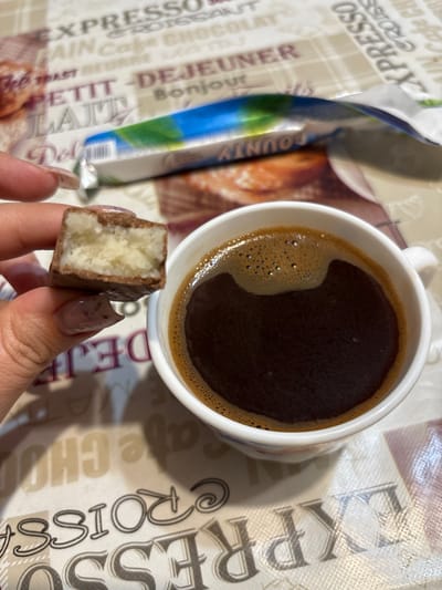 A hand holding a broken chocolate bar is positioned over a white mug of black coffee. The chocolate bar has a white filling. The mug and the chocolate bar rest on a patterned surface with text in French. The pattern includes words like "EXPRESSO," "CROISSANT," and "CHOCOLAT." The scene is brightly lit, suggesting it is indoors during the daytime. There is no information available about the location in the city of Zubov, Russia.