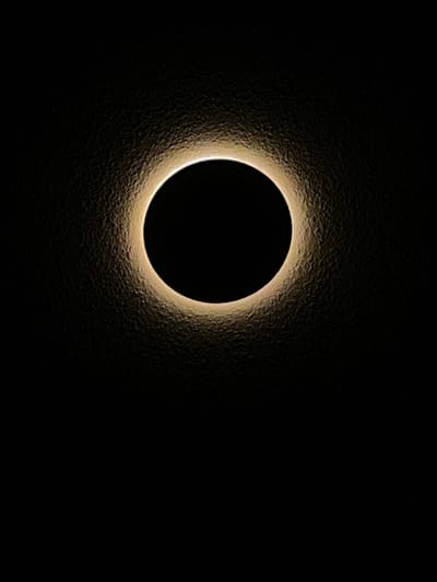The image is a close-up shot of a solar eclipse, focusing on the corona. A perfectly circular, black disc, representing the obscured sun, is centrally located. Surrounding this dark disc is a bright, luminous halo of light, the corona, which flares outward with a textured, irregular edge. The background is entirely black, creating a stark contrast that emphasizes the brilliance of the corona. The texture of the corona suggests a gaseous, ethereal nature. The image captures the intense light and shadow of this celestial event. There are no people, objects, or environmental details indicative of Englewood, United States.