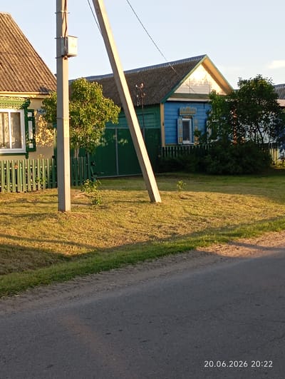 The image captures a rural scene in the Урыцкі сельскі Савет, Belarus, depicting a residential area during daylight. In the foreground, a paved road runs diagonally across the bottom of the frame, with a dirt and grass embankment separating it from a grassy yard. The yard is illuminated by warm sunlight, casting long shadows from a prominent concrete utility pole positioned centrally in the mid-ground.

Behind the utility pole and fence, two houses are visible. The house on the left has a light-colored facade with green trim around a window and a dark, corrugated roof. A green picket fence runs along its front. The house on the right is a striking blue with a dark roof and decorative white and yellow trim around a window. This house is partially obscured by dense green foliage and trees. A green fence also runs in front of this house.

The environment appears to be a typical village setting, with a focus on residential structures and vegetation. The sky is clear and pale blue, indicating fair weather. A timestamp in the bottom right corner reads "20.06.2026 20:22", suggesting the photograph was taken on June 20, 2026, at 8:22 PM. There are no people or animals visible in the image. The overall impression is one of a peaceful, sunlit evening in a Belarusian village.