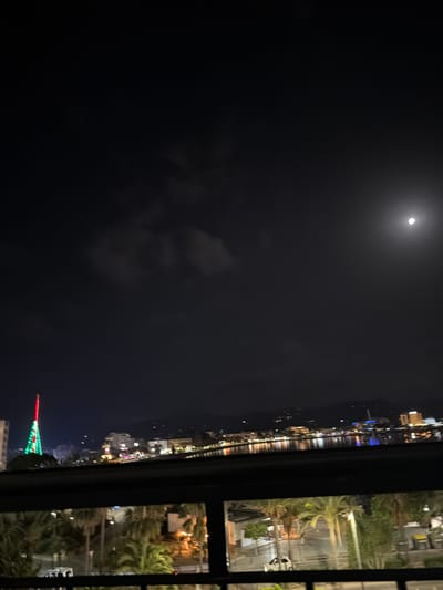 The image captures a nighttime view of the bay in Sant Antoni De Portmany, Spain, as seen from an elevated position. A bright full moon is visible in the dark sky, partially obscured by wispy clouds. In the distance, the city skyline is illuminated with numerous lights reflecting on the calm water of the bay. A prominent, illuminated tower, possibly a landmark or decorative structure, stands out on the left side of the cityscape, adorned with green and red lights. The foreground features a dark, horizontal railing, suggesting the viewpoint is from a balcony or a similar structure. Below the railing, palm trees and some buildings are visible, indicating a coastal urban environment. There are no discernible people or specific actions taking place in the image.