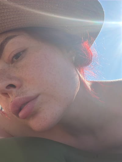 The image is a close-up, low-angle selfie of a person wearing a straw hat and a green garment. The person has fair skin with freckles, reddish-pink hair visible on the right side of their head, and appears to be looking at the camera with their lips slightly parted. The sun is bright and positioned in the upper right corner of the frame, causing lens flare and obscuring parts of the background, which seems to be a clear blue sky. The person's left eye, eyebrow, nose, and mouth are visible. The lighting suggests it is daytime and outdoors. The image does not contain any discernible landmarks or elements that would allow for the identification of a specific location within the city of Узюково, Russia.