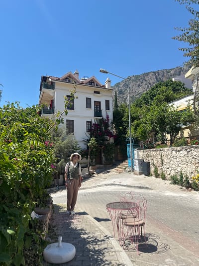 This photograph captures a sunny street scene in Kaş, Turkey, featuring a woman standing on a paved sidewalk. The image, oriented correctly, shows a vibrant outdoor setting under a clear blue sky.

In the foreground, lush green bushes with scattered pink flowers partially frame the lower left. To the right, a white, round umbrella base sits on the sidewalk. Further along the sidewalk, two pink wrought-iron cafe tables and chairs, adorned with ornate scrolling designs, are visible. Strong shadows cast by these objects suggest bright sunlight.

In the midground, a person, likely a woman, is standing on the sidewalk, facing slightly towards the camera. She is wearing a light-colored hat, a vest over a patterned shirt, and light-colored trousers, with a slight smile on her face. Behind her and to the left stands a prominent multi-story white building with brown-shuttered windows. Reddish-purple bougainvillea vines climb part of its facade. A street lamp extends from the left side of the frame.

The background reveals a large, rocky mountain range under a brilliant, cloudless blue sky, indicating the town's mountainous coastal setting. The overall atmosphere is bright and pleasant, with the clear weather and relaxed posture of the person conveying a sense of calm. No visible text is present in the image.