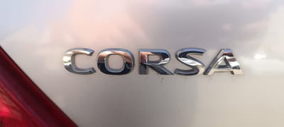 The image displays the chrome-plated word "CORSA" affixed to the rear of a light-colored vehicle. The letters are positioned horizontally and are slightly raised from the car's surface. To the left of the word, a portion of a red taillight is visible. The background is the car's body, which appears to be a pale metallic color, with some reflections of the sky and surroundings visible on its surface. There are a few small specks of dirt or debris on the car's paint around the letters. The photograph was taken outdoors, as indicated by the reflections and lighting. The scene is consistent with the rear of a car parked in a street or parking lot in Schwaikheim, Germany, likely during daylight hours.