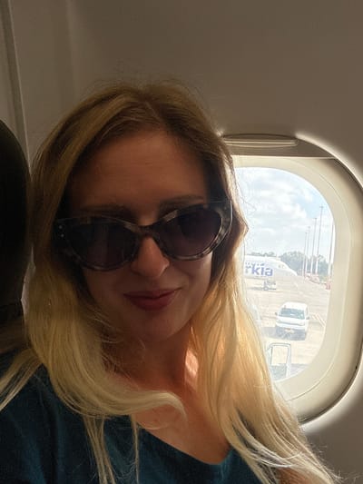 A woman with blonde hair, wearing sunglasses and a dark teal shirt, is seated on an airplane, taking a selfie by a window. Outside the window, an Arkia airplane is parked on the tarmac at Ben Gurion Airport, located in Yehud-Monosson, Israel. The sky is visible and appears to be partly cloudy. Several lights or poles and a white van are also visible on the airport grounds.