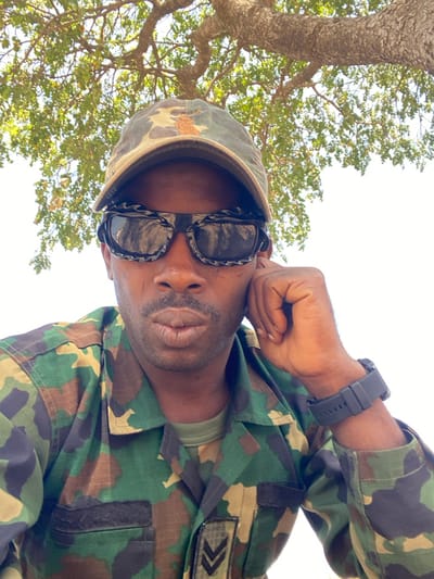 An adult male, located in Zaki Biyem, Nigeria, is depicted in a head-and-shoulders portrait, captured outdoors with an upward angle. He wears a camouflage baseball cap with a light-colored emblem on the front, dark sunglasses featuring a black and white patterned frame, and a camouflage uniform shirt over an olive green undershirt. The uniform shirt displays two black chevron rank insignia on the right chest area. His left hand is raised, with fingers resting against his jawline and ear. He has short facial hair and his lips are slightly pursed while looking directly at the camera. The background consists of tree branches with green leaves against a bright, clear sky. The specific location within Zaki Biyem is not discernible.