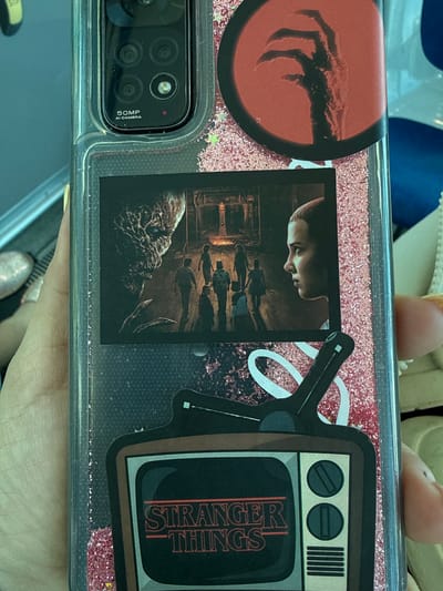 The image shows a close-up of a phone case decorated with graphics related to the TV show "Stranger Things." The phone case has a clear plastic exterior with pink glitter embedded within.
Prominently featured are several stickers:
1. A circular sticker on the left depicts a red, claw-like hand reaching out from a dark red background, a common motif associated with the Demogorgon from the show.
2. A rectangular sticker in the center displays a scene from the show, featuring the character Eleven in profile looking down at a group of shadowy figures in what appears to be an indoor, dimly lit setting.
3. To the right, a sticker designed to look like an old television set features the "Stranger Things" logo in its screen. The TV graphic also has two circular knobs and a set of vertical lines on the top right, representing ventilation.
The camera module of the phone is visible in the lower left corner, indicating the type of phone. The text "50MP AI CAMERA" is visible next to the camera lenses. The phone itself is likely placed on a dark surface, possibly a table or desk.
The overall aesthetic is dark and fantastical, reflecting the themes of "Stranger Things." There are no explicit cues about the time of day, weather, or specific location within Getafe, Spain, beyond the context provided. The image appears to be a static shot of the phone case, with no active event or clear emotional expression depicted.