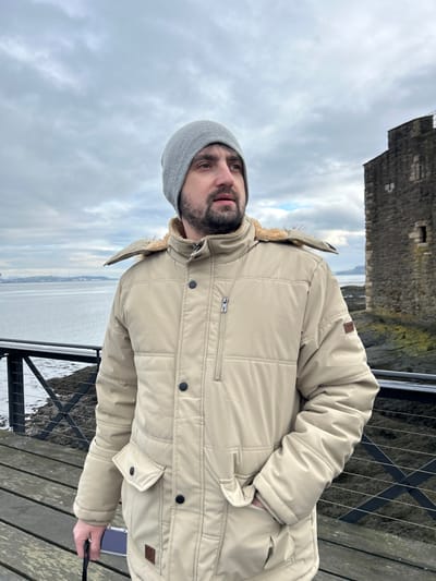 An adult male stands outdoors on a wooden deck in Blackness, United Kingdom, with Blackness Castle visible to his right. He is positioned facing slightly to the viewer's right, gazing towards the left horizon. The man wears a grey knit beanie and a beige, quilted winter coat featuring a faux fur-lined hood, black buttons, zippered chest pocket, lower flap pockets, and side-entry pockets. His left hand is placed inside a coat pocket, while his right hand, down by his side, holds a dark rectangular object, possibly a smartphone or camera. A black metal railing with horizontal and diagonal bars is in the immediate foreground. Beyond the railing lies a body of water, likely the Firth of Forth, with a rocky, moss-covered shoreline directly adjacent to the historic stone structure of Blackness Castle on the right. In the distant background, a faint shoreline or landmass is visible across the water. The sky is overcast with a uniform grey cloud cover.
