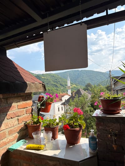 The image depicts a view from a covered balcony overlooking a town nestled in a valley with rolling, tree-covered hills in the background. A minaret of a mosque is visible among the buildings. The foreground shows a tiled surface with several potted geranium plants, some of which are in bloom with bright pink and red flowers. Also on the surface are clear plastic cups, a yellow cloth or sponge, a light blue cleaning cloth, and a bottle of hand soap. A brick wall and a portion of a shingled roof are on the left side of the frame. The sky is partly cloudy and blue. The scene appears to be a residential area, possibly with a cafe or guesthouse given the setup on the balcony. The overall impression is one of a sunny day in a picturesque, possibly historic, European town.