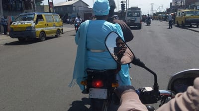 The image shows a person riding a motorcycle in Rubavu, Rwanda, at coordinates -1.664, 29.275. The rider is wearing a light blue headscarf and a light blue dress. The rider is holding onto the handlebars of the motorcycle, and a second person is seated behind the rider. The motorcycle is facing forward, and the rear taillight is visible. There are two other vehicles in the background, both of which are vans. The road is paved and appears to be in good condition. The sky is clear and blue.  The scene is a busy street with people walking and driving.
