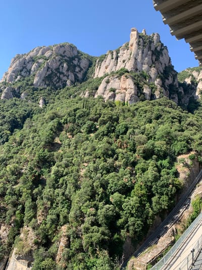 The image depicts a vast, steep mountain covered in dense green foliage. Jagged, light-colored rock formations protrude from the dense vegetation, particularly on the left side of the frame, suggesting a mountainous terrain. A clear, bright blue sky dominates the upper left portion of the image. The perspective is from an elevated viewpoint, looking down and across the mountainside. In the upper right quadrant, a modern bridge or walkway, constructed with metal and glass, stretches across a portion of the mountain. This structure is supported by railings and appears to be a pathway designed to navigate the steep inclines. The scene is bathed in daylight, indicating a sunny day, and the lush greenery suggests a temperate climate. The location is identified as Monistrol de Montserrat, Spain, which is known for its distinctive mountain formations. No people are visible in the image.