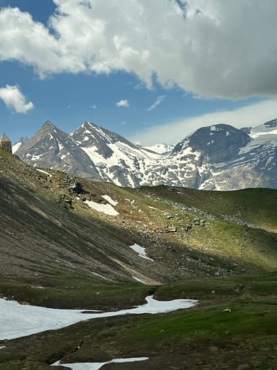 The media file depicts a mountain landscape, likely in Fusch an der Großglocknerstraße, Austria, on a sunny day with scattered clouds. The scene is dominated by a vast mountain range with snow-capped peaks, suggesting a high-altitude location. The foreground features a steep, grassy slope interspersed with patches of lingering snow and rocky outcrops. The colors are vibrant, with lush green vegetation contrasting against the stark white snow and grey rock.

The image is oriented vertically, creating a sense of depth and grandeur. The sky is a bright blue, partially obscured by fluffy white clouds. The sunlight casts shadows on the mountainside, highlighting the rugged terrain. The overall impression is one of natural beauty and vastness, characteristic of alpine environments. There are no people or man-made objects visible in the scene, emphasizing the wild and untouched nature of the landscape. The time of day appears to be midday or early afternoon, given the bright sunlight and clear sky. The weather is pleasant and conducive to outdoor exploration.