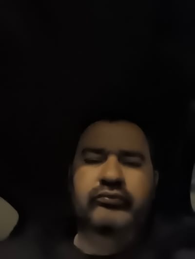 A dimly lit, low-resolution image depicts an adult male from the chest up, seemingly positioned within a vehicle interior or dark room. The subject has a dark complexion, a receding hairline or closely shaven head, and prominent dark facial hair consisting of a mustache and beard. His eyes appear narrowed or partially closed, and his mouth is slightly open. He is dressed in dark clothing. The background is predominantly black, with a lighter, beige or tan-colored object partially visible on the left side of the frame and a subtle light reflection on the far right. No specific actions or interactions are discernible. The scene is described as taking place in Rosolini, Italy.