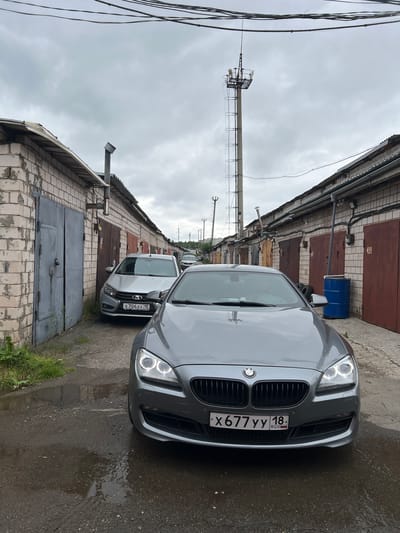 The image shows a row of brick garages in the city of Veteran-2, Russia, on an overcast day. In the foreground, a grey BMW coupe is parked. Behind it, a silver Lada sedan is parked next to a large grey garage door. Further down the row of garages, other cars and a blue barrel are visible. A utility pole with antennas stands tall in the background. The ground is wet, suggesting recent rain.