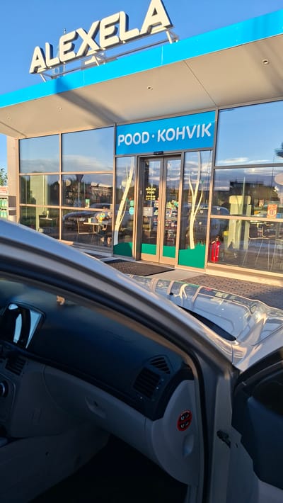 The image shows a view from inside a car parked in front of a ALEXELA gas station and convenience store in Tiskre, Estonia. The gas station has a prominent blue and white sign that says "ALEXELA". Below the sign, on the front of the building, is a blue banner with white text that reads "POOD · KOHVIK", indicating it is a shop and cafe. The building features large glass windows and double glass doors. Inside the store, shelves stocked with various products are visible, along with tables and chairs set up for customers. Outside the store, a red fire extinguisher is visible next to the entrance. The car interior in the foreground shows the dashboard, steering wheel, and a side door ajar, with an "AIRBAG" warning sticker visible on the interior panel. The sky is clear and blue, suggesting it is daytime.