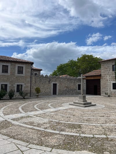 The image shows a stone square in Ávila, Spain. The square is paved with small cobblestones arranged in a pattern of concentric circles around a central stone cross. Buildings surround the square, constructed of grey stone with dark windows and red tile roofs. A green tree stands behind one of the buildings. The sky above is blue with white clouds.