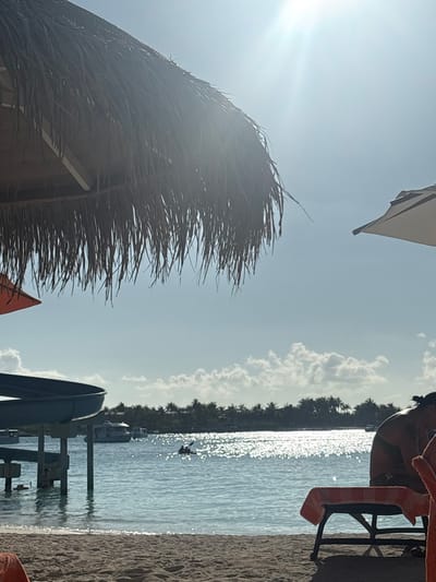This image depicts a sunny beach scene in Himmafushi, Maldives. In the immediate foreground, sandy beach occupies the lower portion of the frame. Dominating the upper left, a large thatched umbrella casts shade, with a portion of an orange fabric umbrella visible underneath. To the right, another light-colored fabric umbrella is partially visible against the sky. A person with dark hair pulled back is seated on a beach lounge chair, partially obscured on the right, covered by an orange-striped towel.
In the midground, the clear, light blue ocean water glistens with strong sun reflections across its surface. The supports of a blue waterslide extend into the water from the left. A small vessel, possibly a kayak, containing one individual is present in the open water. Several larger boats are anchored further offshore.
The background features a distinct landmass, likely an island, densely covered with green palm trees. The sky above is clear blue with scattered white cumulus clouds. A bright sun flare is prominent in the upper center of the image.