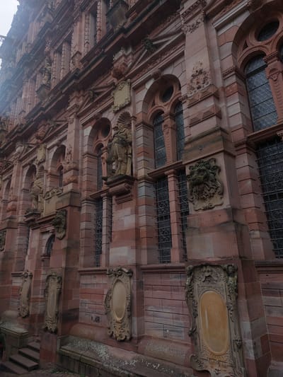 The image shows a close-up, eye-level, outdoor shot of the elaborate stone facade of Heidelberg Castle in Heidelberg, Germany. The architecture is primarily red sandstone with intricate carvings and architectural details, including arched windows with dark metal grates, stone sculptures of figures in armor, and decorative reliefs of grotesque faces and ornamental motifs. The lighting suggests an overcast day. The lower portion of the image shows a stone staircase leading up to a lower level of the castle and a stone bench-like structure with mossy growth. The overall impression is one of historical grandeur and weathered beauty.