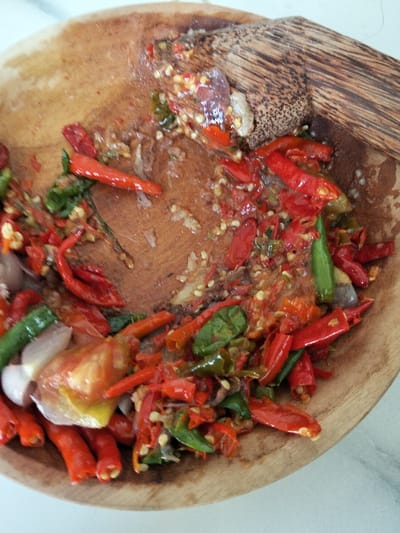 A wooden mortar holds a mix of smashed ingredients for chili sauce. Red and green chili peppers, along with pieces of onion and tomato, are mashed together. Some ingredients are coarsely chopped, while others are finely ground into a paste. A wooden pestle rests on the side, suggesting the ongoing preparation of the sauce. The bowl shows signs of wear, indicating frequent use.