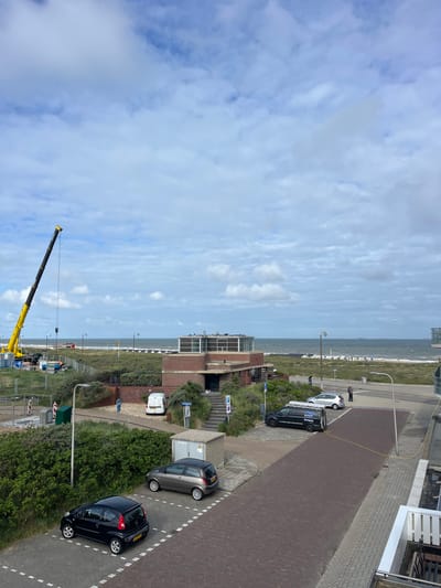 This image captures a coastal scene in Katwijk, Netherlands, viewed from an elevated vantage point, likely a building, looking towards the North Sea. The sky is predominantly light blue with scattered white and grey clouds, indicating an overcast or partly cloudy daytime.

The horizon shows the North Sea, with several distant ships or boats. A sandy beach is visible along the coastline, dotted with a few beach chairs and umbrellas. Inland from the beach, there's a strip of green, bushy vegetation, characteristic of dunes or coastal landscaping, separating the beach from roads and structures.

Paved roads and walkways are prominent, featuring dashed white lines and some yellow markings. Various vehicles are parked along these roads: a dark van with "KOOIJ Reiniging" (Dutch for cleaning) text on its side and ladders on its roof, a silver hatchback parked next to it, and further along, a dark grey hatchback and a black hatchback. A white van is partially visible near a building, and a black SUV is parked closer to the beach area.

Several modern buildings with brick and glass facades are integrated into the scene. Streetlights, trash bins, and signs are visible, including a blue sign with a white wheelchair symbol indicating disabled parking. A large yellow and black mobile crane is partially visible in the lower left portion of the image, with its boom extending upwards into the sky. A few small human figures can be seen, including one person standing near the silver car and others on the beach. The scene depicts a calm day in a coastal town.