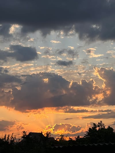 A dark, stormy sky dominates the upper portion of the image, with lighter, scattered clouds below tinged with orange and yellow hues from the setting sun. The sun's rays are visible, bursting through gaps in the clouds, creating a dramatic effect. Silhouetted against the bright sky are the rooftops of houses and trees, suggesting a rural or suburban setting. The overall mood is one of dramatic natural beauty, with the contrast between the dark clouds and the bright sunlight creating a sense of impending change. The location is identified as Slobozia, Moldova.