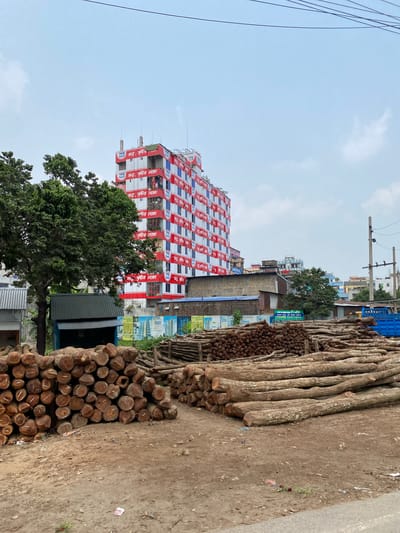 The image shows a pile of logs and tree trunks stacked on a dirt ground next to a road or sidewalk, in what appears to be a city or town. In the background is a tall building, approximately 10 stories high, decorated with red and white banners or signage. Text is visible on the banners, including "জম্ম, সৃষ্টির লক্ষ্যে" in Bangla (Bengali). A large tree is visible in front of the building, partially obscuring it. Other buildings and structures are visible in the background, including smaller shops and residential buildings. Power lines are overhead. The sky is light blue with some clouds. The photograph was taken in Narayanganj, Bangladesh.
