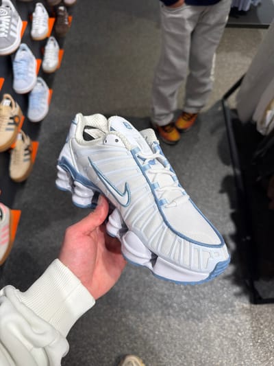 A white and light blue Nike Shox sneaker is held in a person's hand, displayed indoors with other sneakers laid out on a black surface. The setting appears to be a retail store. A person wearing gray pants and orange shoes stands blurred in the background. This image may have been taken in a store in Spreitenbach, Switzerland.