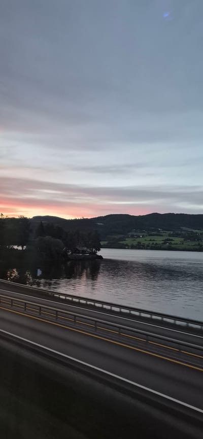 This is a landscape photograph taken at dusk, likely from a bridge or elevated vantage point, overlooking a body of water and a forested hillside. The sky is a gradient of soft grays, pinks, and oranges, suggesting the end of a sunset. Streaks of clouds are visible, adding texture to the sky.

The scene includes a dark, calm body of water reflecting the colors of the sky and the silhouette of the landscape. On the far side of the water, a steep, tree-covered hill rises, with scattered lights and structures suggesting a rural settlement or scattered houses. The greenery of the hillsides is dark due to the low light.

In the foreground, a dark asphalt road with yellow and white lane markings runs diagonally across the frame, indicating a roadway, possibly a highway. A metal guardrail is visible along the edge of the road, bordering the water. The overall mood is serene and tranquil, capturing the quiet beauty of twilight in a natural setting. The location cues, such as the distinct hillside and water, along with the mention of Biri, Norway, suggest a fjord or a large lake in a mountainous or hilly region. No people or specific objects of note are clearly discernible beyond the road, guardrail, and the natural landscape elements. The time of day is clearly evening, as indicated by the sunset colors in the sky and the dim lighting. The weather appears to be clear, allowing for visibility of the sunset.