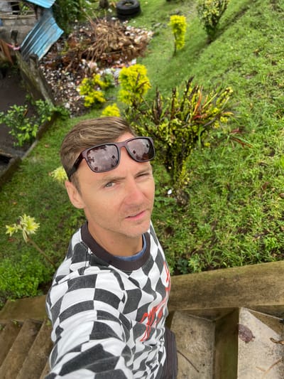 A man is taking a selfie on a set of outdoor concrete stairs. He is wearing sunglasses and a black and white checkered long-sleeved shirt. The background shows a steep, grassy slope with some tropical plants and what appears to be a pile of garbage and debris near a blue metal structure. The setting is likely a residential area or an area with less-developed infrastructure in Klungkung, Indonesia.