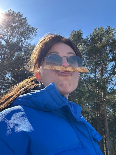 A woman is holding a breadstick between her nose and upper lip. She is wearing sunglasses and a blue puffer jacket. The background shows a clear blue sky, trees, and sunlight. The location is possibly the city of Zīvartiņš, Latvia.