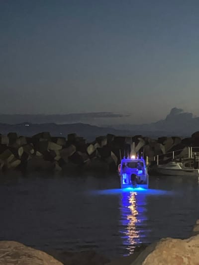 The image captures a nighttime scene within a harbor or marina area in Bellambi, Australia. A white recreational boat is prominently featured, moored in dark water and illuminated by bright blue underwater lights, which cast a vibrant blue reflection directly beneath the vessel. A white stern light is also visible on the boat's upper structure. A single human figure is discernible standing on the boat's rear deck. To the left and behind the boat, a substantial breakwater, constructed from dark, angular concrete tetrapods or large rocks, forms a barrier. To the right of the illuminated boat, a second, smaller, lighter-colored vessel is partially visible, appearing to be docked alongside a pier or similar structure. The background consists of dark, distant mountains or hills under a deep blue-grey sky with scattered clouds, and faint artificial lights are visible on the distant landmass. The immediate foreground features a light-colored, rocky or concrete surface, suggesting the viewpoint from the shore or an adjacent embankment.